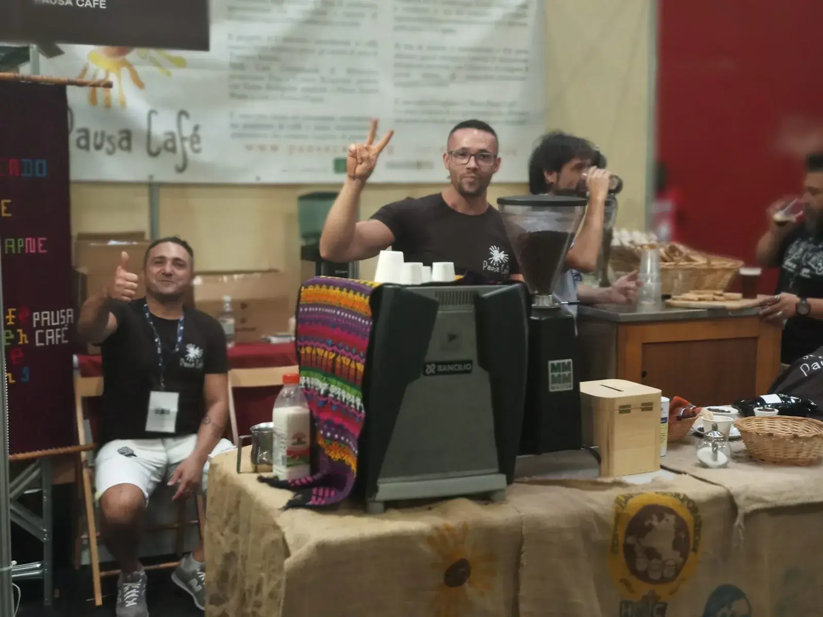 Deux hommes animent un stand de café avec une machine à moudre, l’un faisant un signe de victoire.