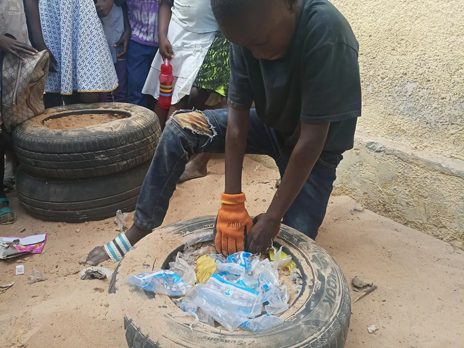 Un jeune remplit de déchets plastiques et de terre un pneu posé au sol.