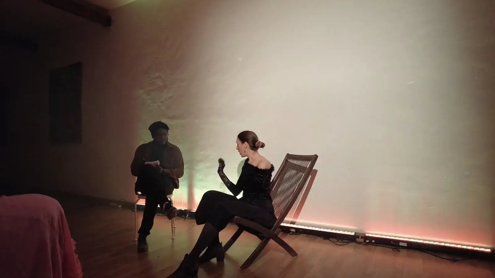Un comédien et une comédienne jouent une scène avec des chaises devant un mur éclairé.