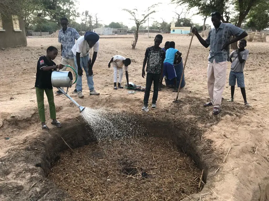 Des enfants arrosent un composteur enterré rempli de paille dans une cour scolaire