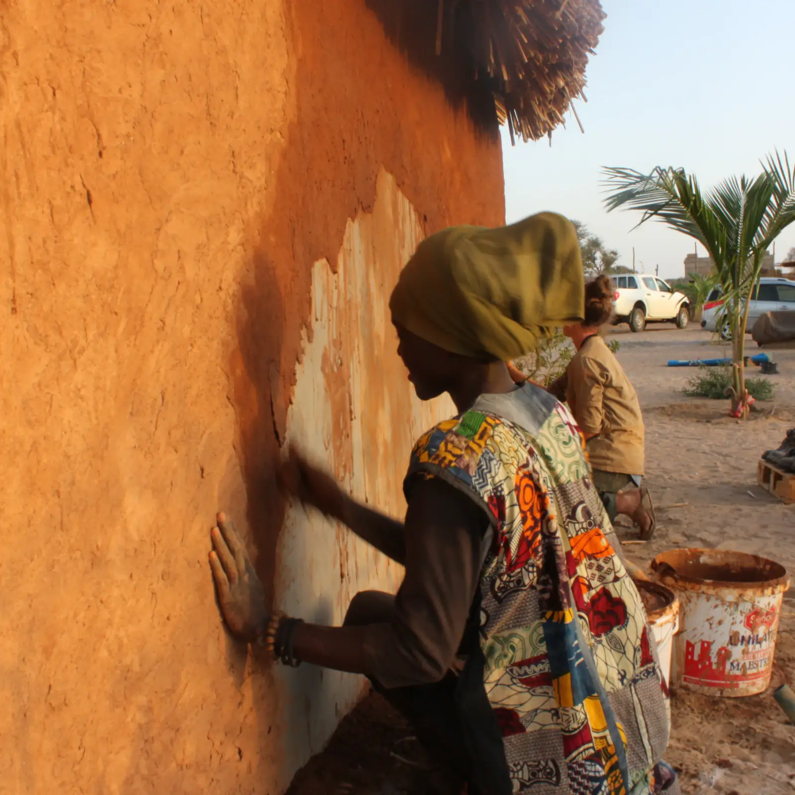 Une femme enduit de terre ocre le mur d’une construction.