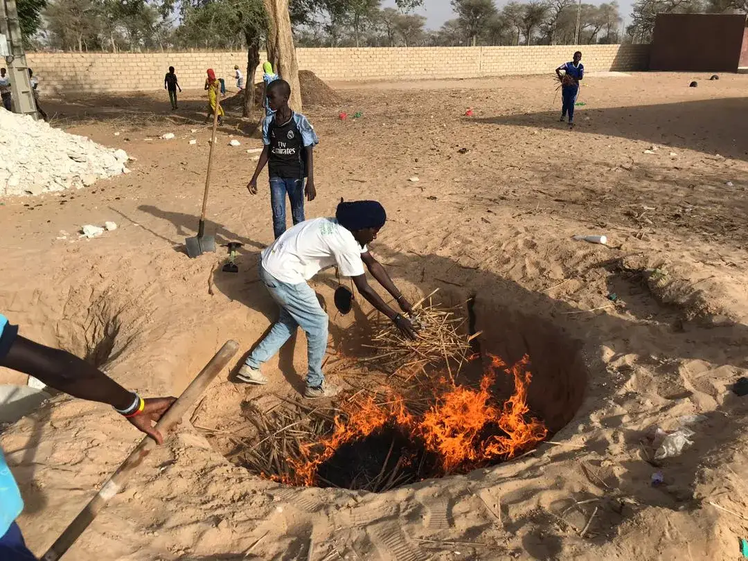 Un homme alimente un feu de branchages dans une fosse, entouré d’enfants dans une cour d’école