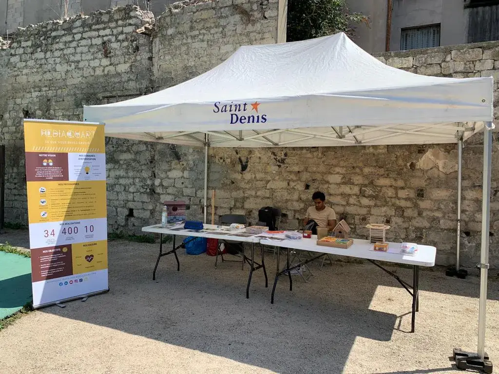 Un stand installé sous une tente blanche devant un mur en pierre, avec un panneau « Sable Denis » et des personnes à table.