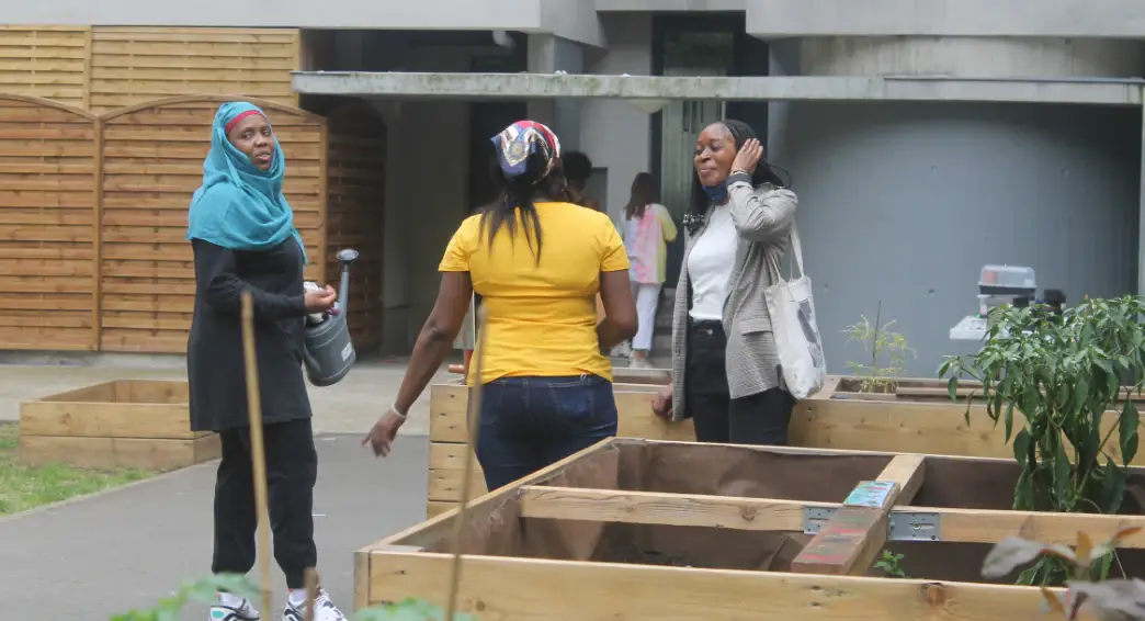 Trois femmes debout près de bacs de culture en bois dans une cour, l’une tenant une pelle.