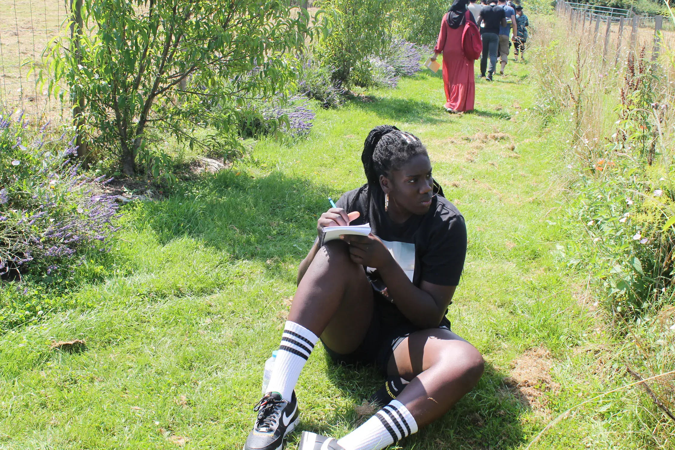 Une jeune femme assise dans l’herbe au bord d’un chemin, un groupe s’éloigne dans le jardin.