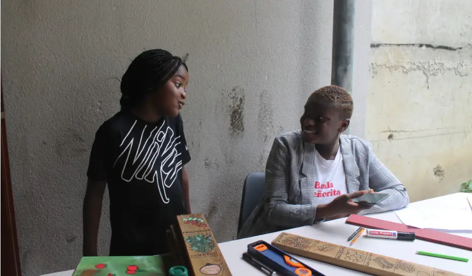 Deux femmes assises à une table couverte de pièces de bois et d’outils, en pleine discussion.