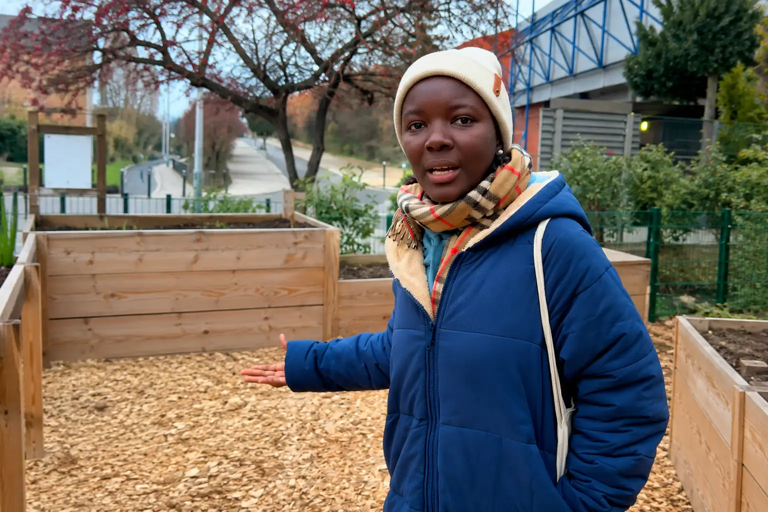 Une enfant en manteau bleu se tient dans un bac de copeaux de bois, devant un jardin partagé.