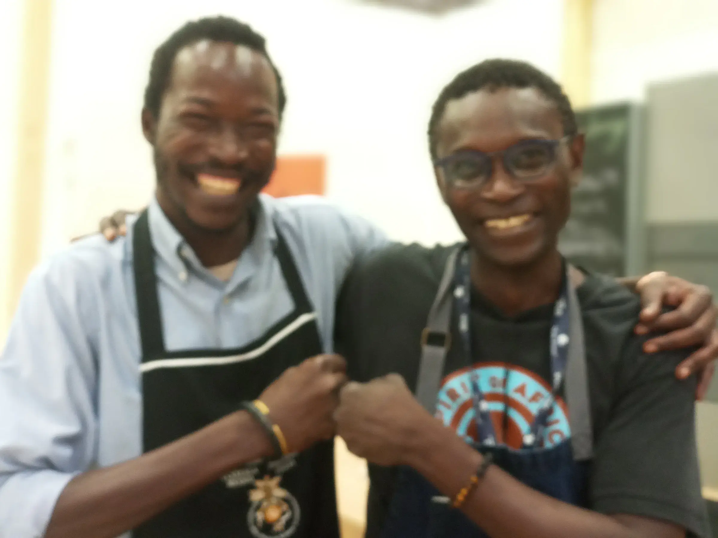Deux hommes souriants en tablier, côte à côte dans un espace de cuisine.