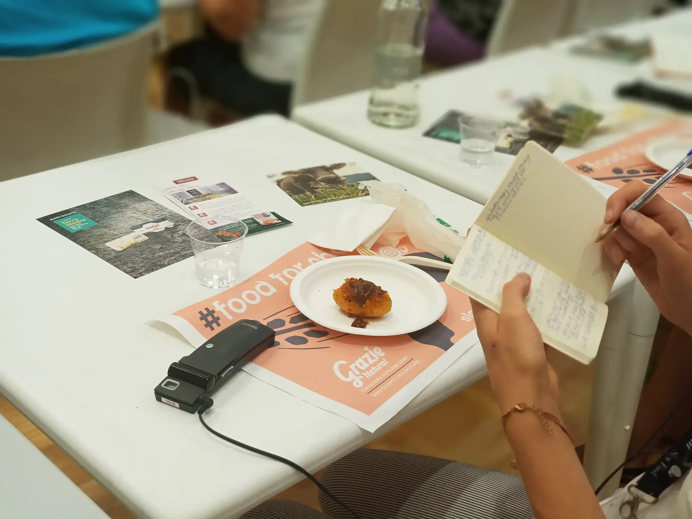 Documents et téléphone posés sur une table, une personne écrit à côté d’une assiette.