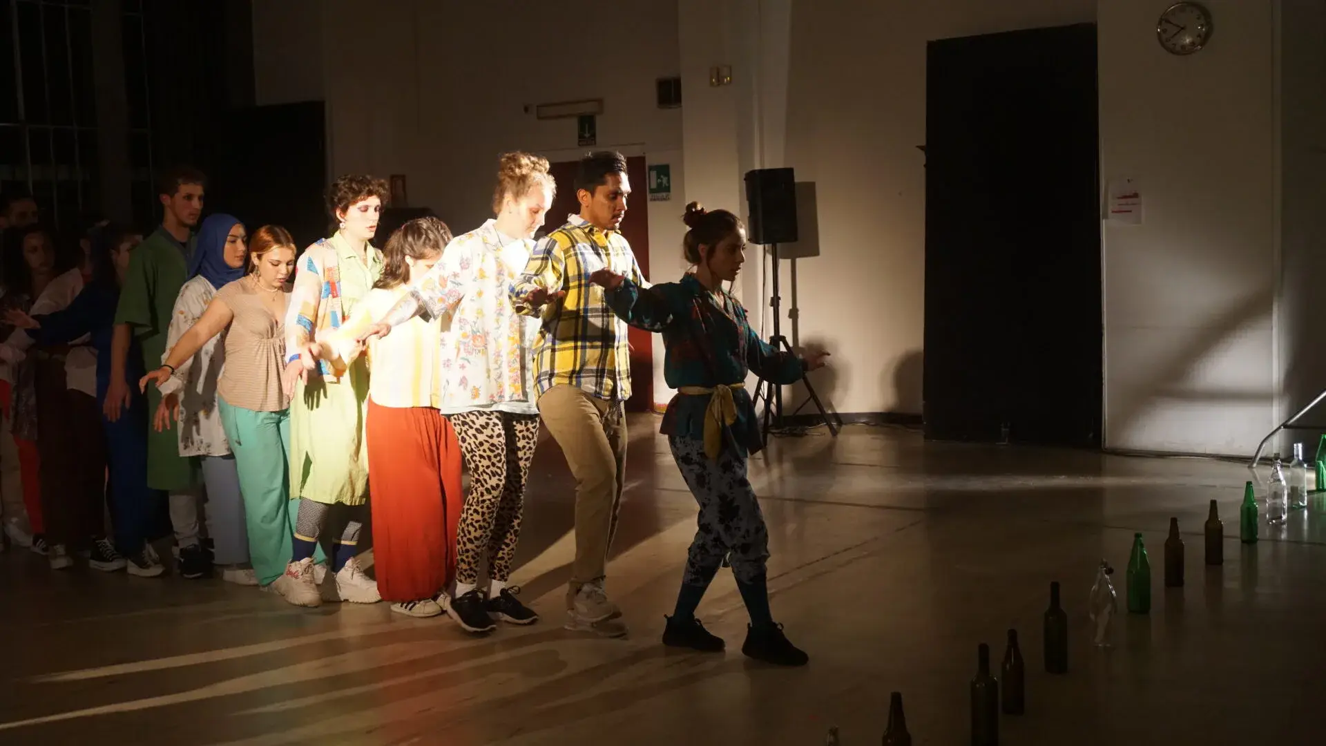 Un groupe de jeunes comédiens sur scène pendant un exercice de théâtre, bouteilles alignées au sol