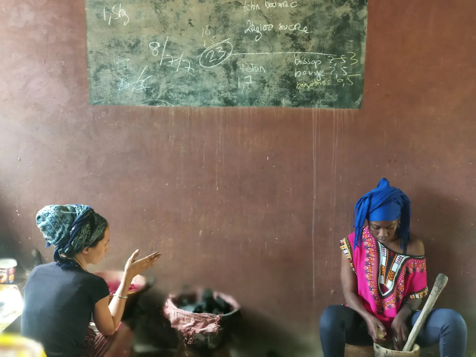 Deux femmes échangent autour d’un mortier dans une pièce, devant un tableau noir